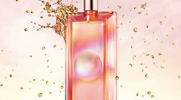 A sleek, rectangular perfume bottle with a metallic cap, featuring a gradient design in shades of pink and orange. Behind the bottle, droplets and splashes of liquid create a dynamic background.