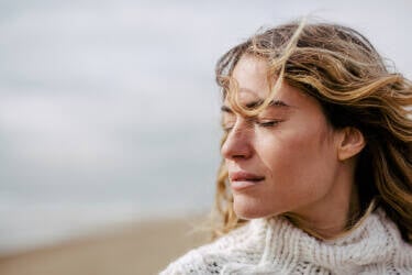 A woman with long, wavy hair stands outdoors with her eyes closed, looking serene against a blurred beach background. She is wearing a cozy, knitted white sweater.