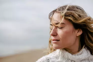 A woman with long, wavy hair stands outdoors with her eyes closed, looking serene against a blurred beach background. She is wearing a cozy, knitted white sweater.