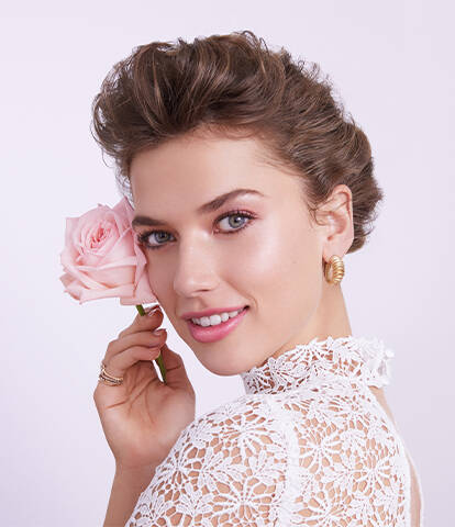 A woman in a lace dress holds a pink rose near her face, smiling softly against a blurred pastel background.