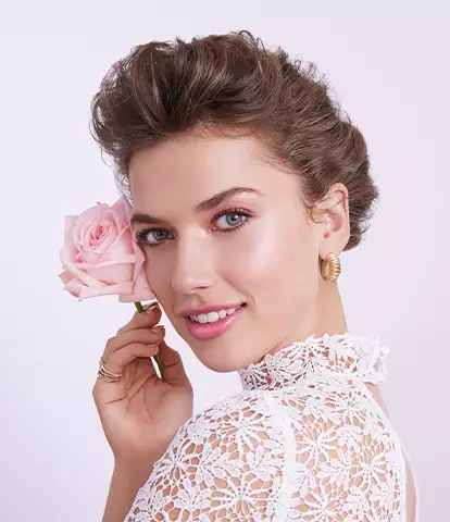 A woman in a lace dress holds a pink rose near her face, smiling softly against a blurred pastel background.