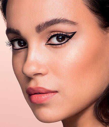 A close-up profile of a woman with striking winged eyeliner and glowing skin, set against a soft pink background.
