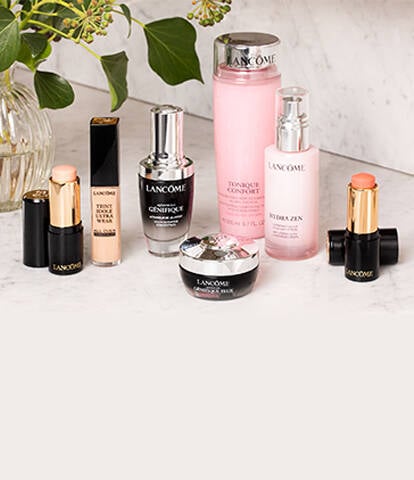 A collection of Lancome skincare and makeup products displayed on a marble surface, including serums, moisturizers, and lipsticks, alongside a vase with greenery.
