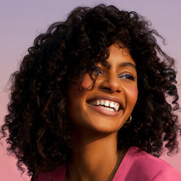 A smiling woman with curly hair, wearing a pink top, against a soft, pastel-colored background.