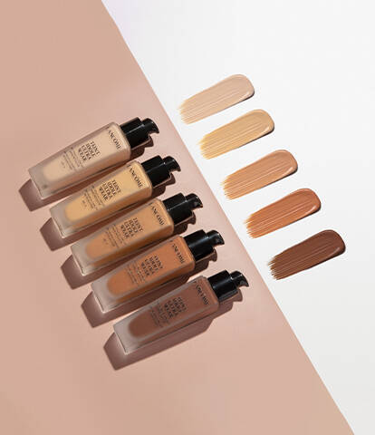A collection of six foundation bottles arranged in a line, with swatches of different skin tones displayed above them on a gradient background.