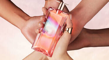 A stylish, colorful perfume bottle held by multiple hands, showcasing a vibrant design against a soft, pastel background.