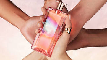 A stylish, colorful perfume bottle held by multiple hands, showcasing a vibrant design against a soft, pastel background.