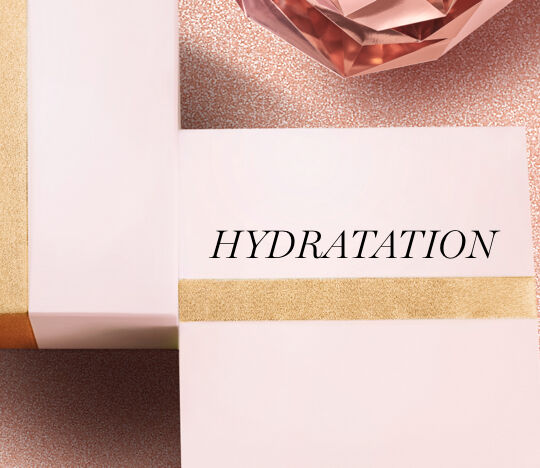 Two elegant boxes with a soft pink exterior, one wrapped in a gold ribbon, featuring the word "HYDRATATION" prominently displayed. In the background, a rose-colored, crystal-like object adds a touch of glamour against a textured, light background.