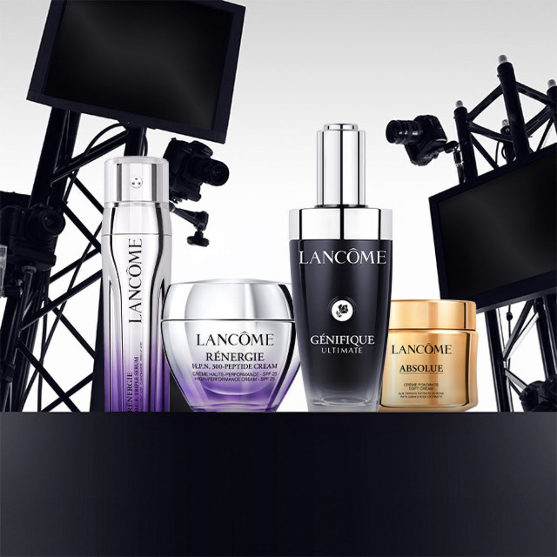 An arrangement of four Lancome skincare products displayed on a sleek black surface, with blurred backdrop elements resembling studio lighting equipment. The products include a serum and three cream jars.