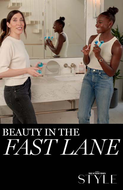 A smiling woman holding beauty products stands next to another woman who is engaging with her in a stylish bathroom setting. The text overlay reads "Beauty in the Fast Lane."