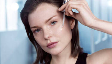 A woman with clear skin and light makeup applies a drop of serum to her face using a pipette, against a blurred blue background.