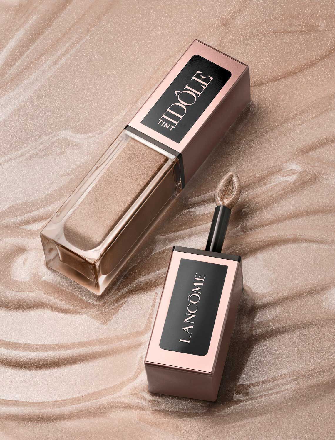 A tube of Lancome's Idole Tint in a sleek bottle, lying on a shimmering gold surface. The applicator is featured alongside the tube, emphasizing its smooth texture.