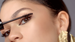 A close-up of a woman's eye applying mascara, showcasing long, defined eyelashes and a bold eyeliner look, with her hair pulled back and wearing statement earrings.