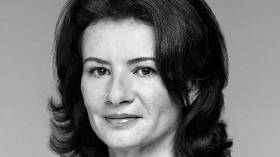 A close-up portrait of a woman with dark, wavy hair and a confident expression, set against a neutral background.