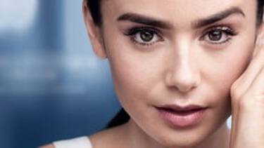 A close-up of a woman with dark hair and natural makeup, looking at the viewer with her hand near her face, against a blurred blue background.