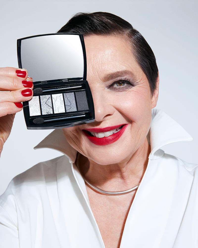 A smiling older woman with short dark hair and bright red lipstick holds a compact eyeshadow palette up to her face, showcasing shades of gray and silver. She wears a crisp white blouse with a folded collar.