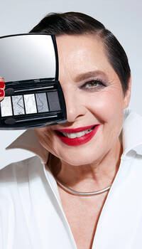 A smiling older woman with short dark hair and bright red lipstick holds a compact eyeshadow palette up to her face, showcasing shades of gray and silver. She wears a crisp white blouse with a folded collar.