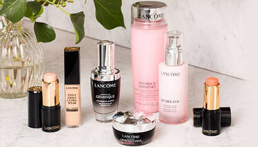 A variety of Lancome beauty products, including foundation sticks, serums, toners, and moisturizers, are arranged on a white marble surface next to a glass vase with greenery.