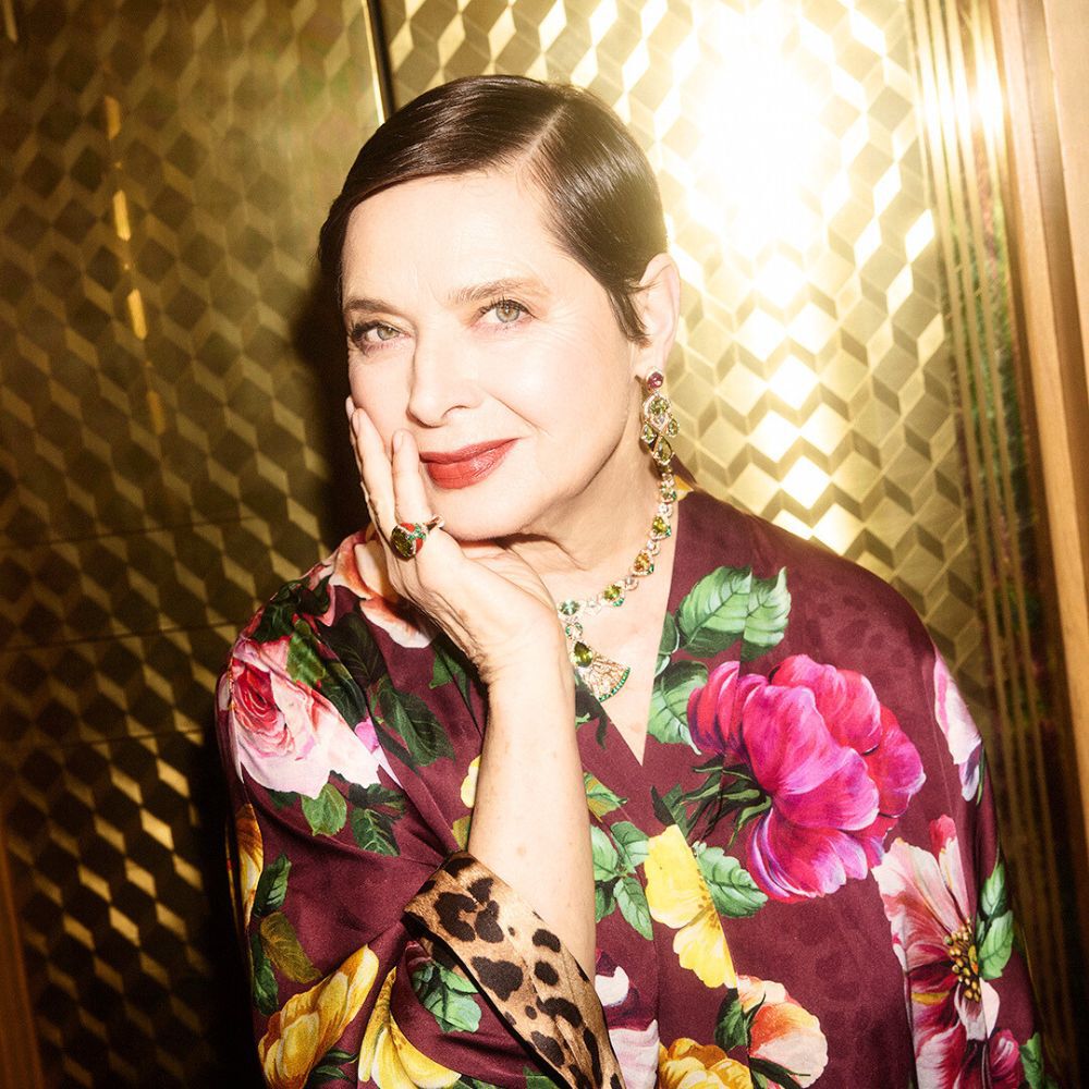 A woman with short black hair and a floral robe poses elegantly, resting her chin on her hand, surrounded by a softly lit golden background. She wears statement jewelry that complements her sophisticated look.