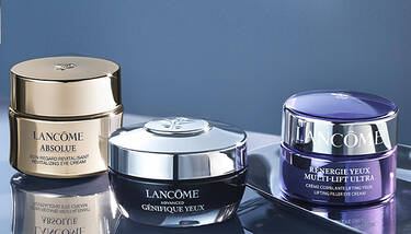 Three Lancome eye creams—Absolue, Genifique, and Renergie Multi-Lift Ultra—are arranged on a sleek, reflective surface against a dark blue background.