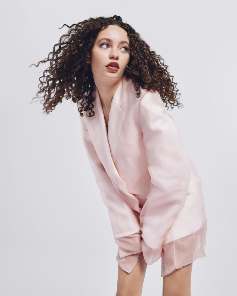 A model with curly hair wearing a light pink oversized blazer poses against a plain background, showcasing a confident expression.