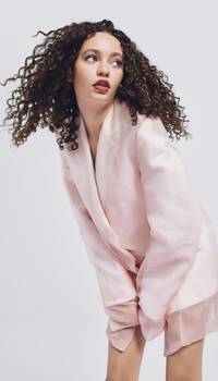 A model with curly hair wearing a light pink oversized blazer poses against a plain background, showcasing a confident expression.