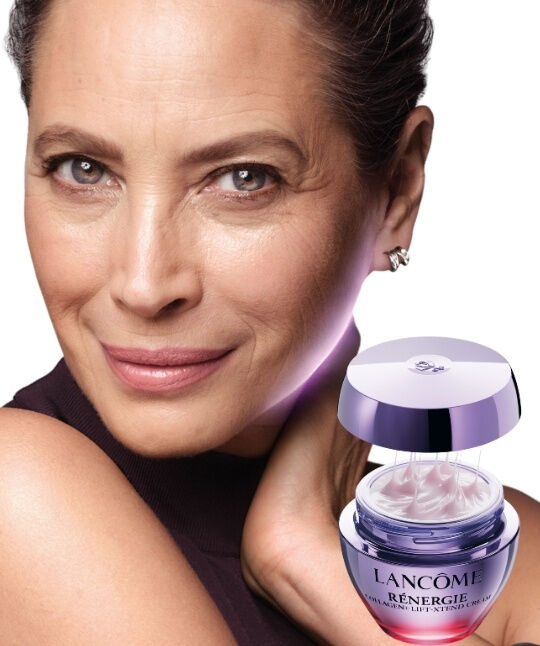 Three spray bottles of Lancome "La vie est belle" Hair & Body Mist in different shades of pink and orange, set against a background of raspberries and a white flower.