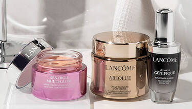 Three Lancome skincare products: Renergie Multi-Glow, Absolue Soft Cream, and Advanced Genifique concentrate, are arranged on a white surface with a blurry background.