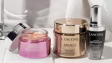 Three Lancome skincare products: Renergie Multi-Glow, Absolue Soft Cream, and Advanced Genifique concentrate, are arranged on a white surface with a blurry background.