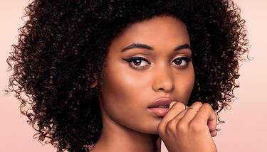 A woman with voluminous curly hair and striking winged eyeliner gazes forward, her finger gently touching her lips, against a soft pink background.