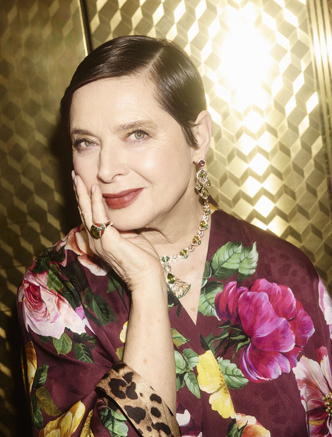 A woman with sleek, short hair poses gracefully, resting her chin on her hand. She wears a floral robe and elegant jewelry, smiling softly against a shimmering gold background.