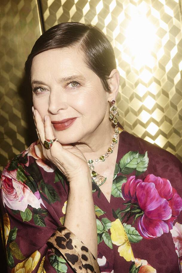 A woman with sleek, short hair poses gracefully, resting her chin on her hand. She wears a floral robe and elegant jewelry, smiling softly against a shimmering gold background.