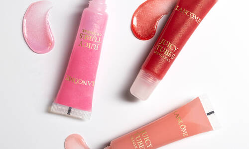 Three tubes of Lancome Juicy Tubes lip gloss in pink, red, and peach shades, with corresponding swirls of gloss beside them.