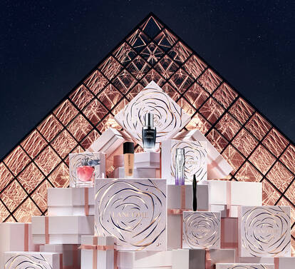A model in a sleek white dress poses next to elegantly stacked gift boxes in front of the Louvre Pyramid at night. The scene is illuminated, highlighting the modern and artistic ambiance.