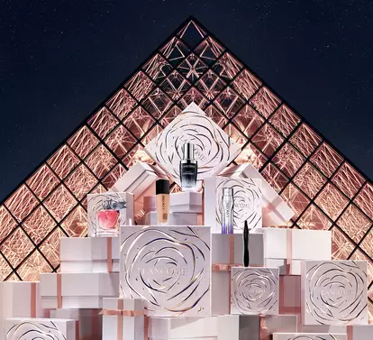 A model in a sleek white dress poses next to elegantly stacked gift boxes in front of the Louvre Pyramid at night. The scene is illuminated, highlighting the modern and artistic ambiance.