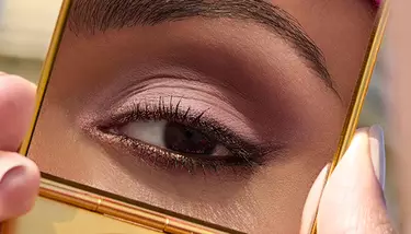 A close-up of an eye with subtle pink and brown eyeshadow, dark eyeliner, and mascara, reflected in a gold compact mirror held by manicured fingers.
