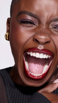 A close-up of a person with short hair, wearing bold red lipstick, smiling widely and laughing, showcasing expressive eyes and ear jewelry against a plain background.