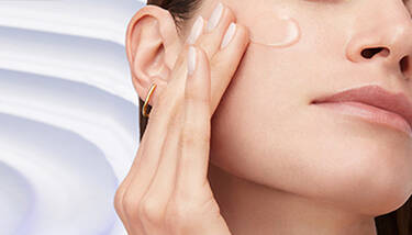 A woman with clear skin and a gold hoop earring gently touches her cheek, where a drop of translucent product is visible, against a soft, light background.