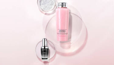 Two Lancome skincare products, Tonique Confort and Advanced Genifique, are elegantly presented on a pink and white background with translucent circular elements.