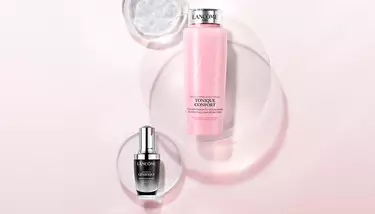 Two Lancome skincare products, Tonique Confort and Advanced Genifique, are elegantly presented on a pink and white background with translucent circular elements.