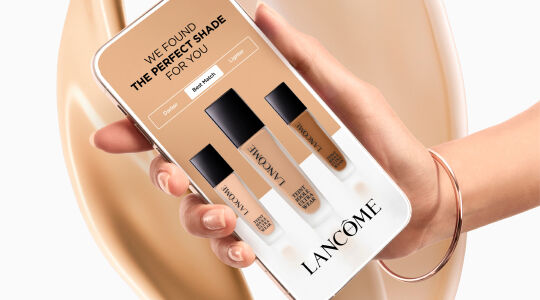 A hand holding a smartphone displaying a Lancome makeup shade finder, with three foundation bottles labeled in shades like "Honey" set against a backdrop of flowing liquid foundation.