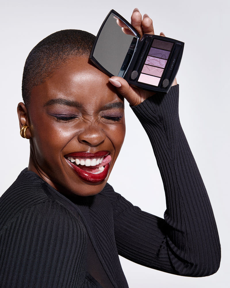 A smiling woman with short hair holds an eyeshadow palette up to her face, showcasing vibrant eye colors. She is wearing a black ribbed top and has bold red lipstick on.