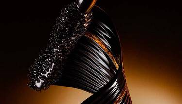 A close-up shot of a mascara wand coated in black product, alongside a sleek, twisted black and gold element, set against a dark, warm-toned background.