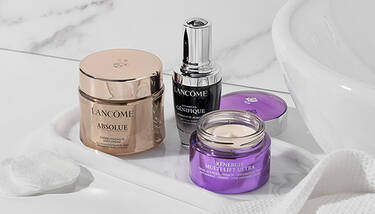 Lancome Absolue Soft Cream, Advanced Genifique serum, and Renergie Multi-Lift Ultra cream are displayed on a white tray with cotton pads, set against a marble background with a white sink.