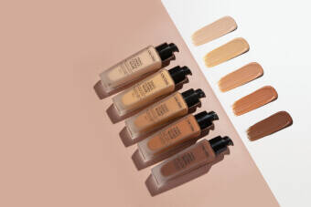 Five foundation bottles in varying shades are arranged on a soft, neutral background, with corresponding swatches of each shade displayed above them.