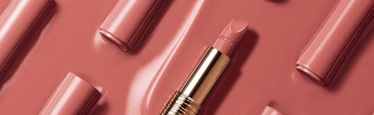 A close-up of a nude pink lipstick tube lying on a glossy pink background, surrounded by similar lipstick tubes arranged neatly.
