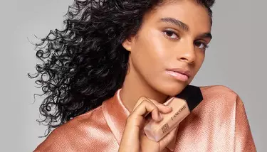A woman with dark curly hair and a subtle glow on her face is holding a Lancome Teint Idole Ultra Wear foundation bottle, against a plain gray background.
