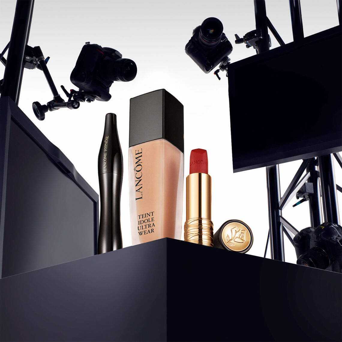 A collection of Lancome makeup products, including a foundation bottle, a mascara tube, a red lipstick, and a compact, is displayed on a dark surface with cameras and lights in the background.