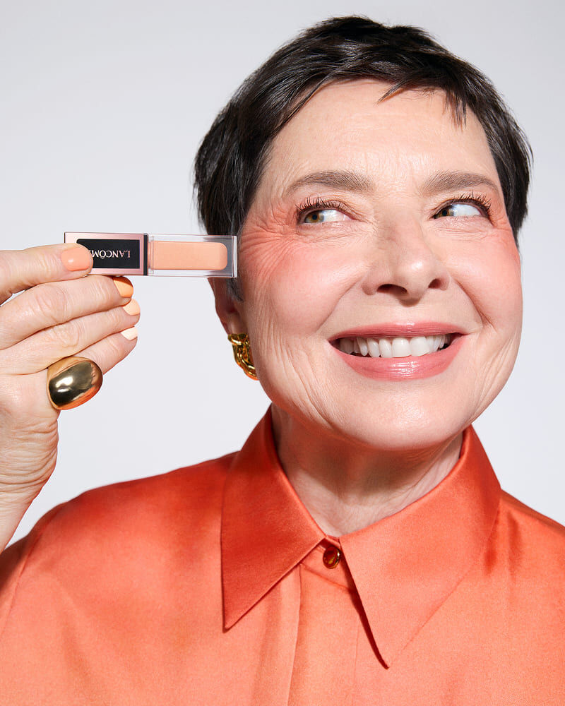 A smiling older woman with short hair holds a small makeup product up to her eye, wearing an orange blouse and gold jewelry.