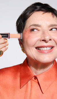 A smiling older woman with short hair holds a small makeup product up to her eye, wearing an orange blouse and gold jewelry.
